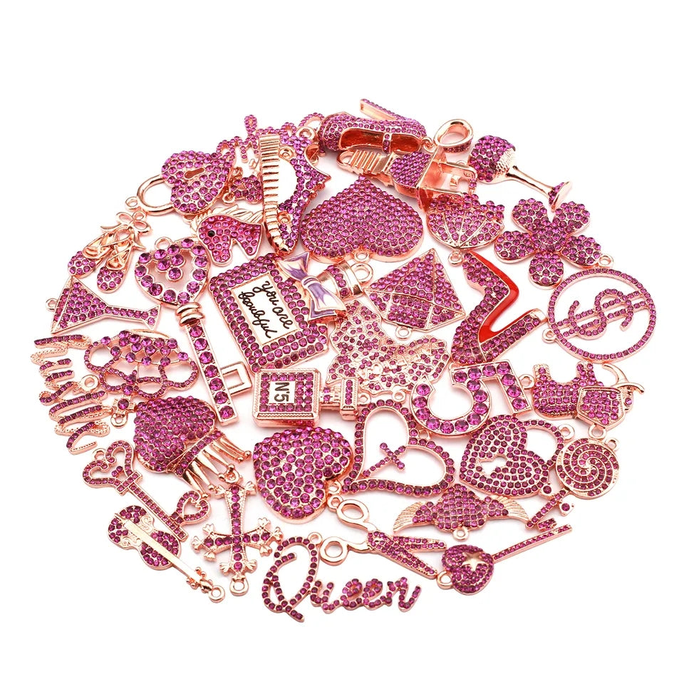 pink rhinestone-embellished charms and jewelry pieces on a plain white background, including hearts, money symbols, a key, a lipstick case, a dollar sign, and a script charm reading &