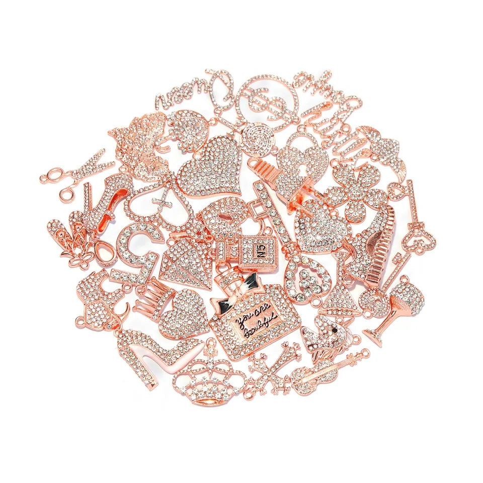 A pile of rose gold rhinestone charms and jewelry findings on a white background, including high heels, hearts, padlocks, a perfume bottle, and letters. $1.88ea